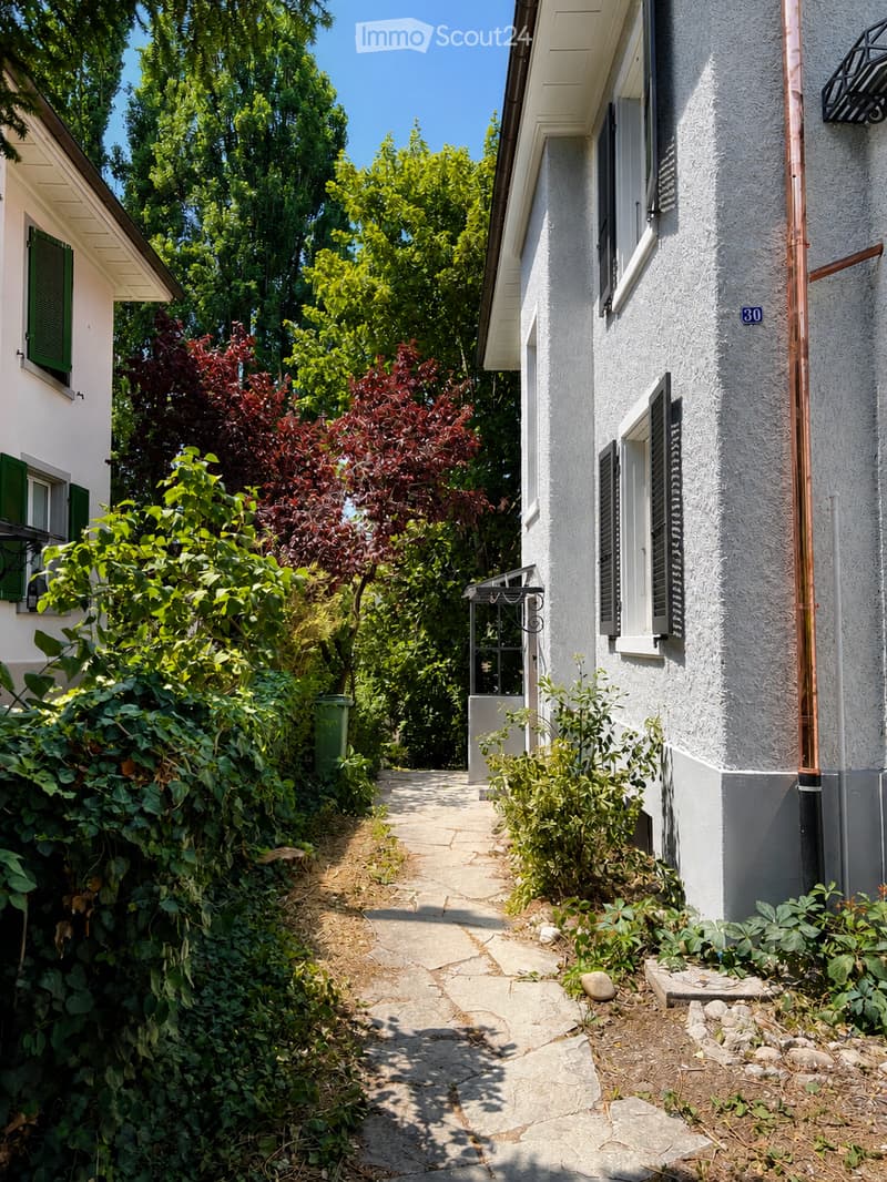 A narrow garden pathway lined with greenery leads to a white house with green shutters and a small balcony. The house has a textured facade and a visible address number30.