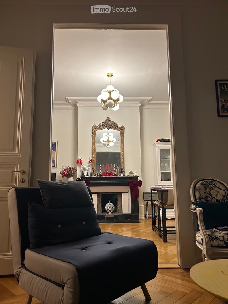 Elegant living room with a dark blue sofa, floral-patterned armchair, small round table, ornate mirror above fireplace, and decorative items on mantel.