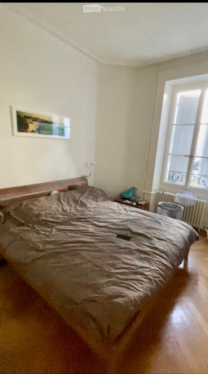 Bedroom with a large bed, nightstand, picture frame on the wall, radiator, and a trash bin near the window.