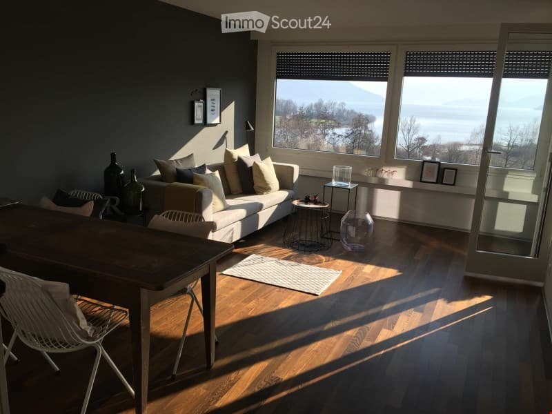 Bright living room with large windows offering a scenic view, featuring a white sofa with multiple cushions, a wooden coffee table, a dining table with chairs, and decorative vases.