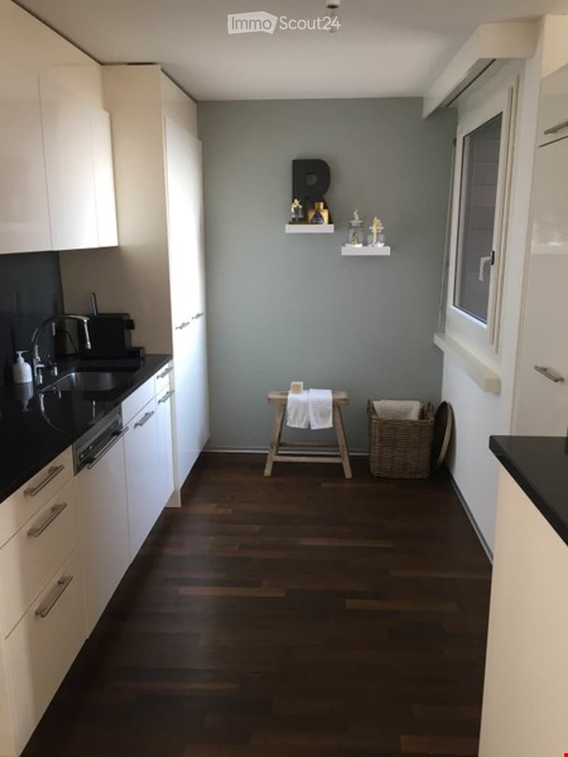 Modern kitchen with white cabinets, black countertops, and a wooden floor. Features a sink, faucet, dishwasher, small wooden stool, woven basket, and decorative shelves with candles and figurines.