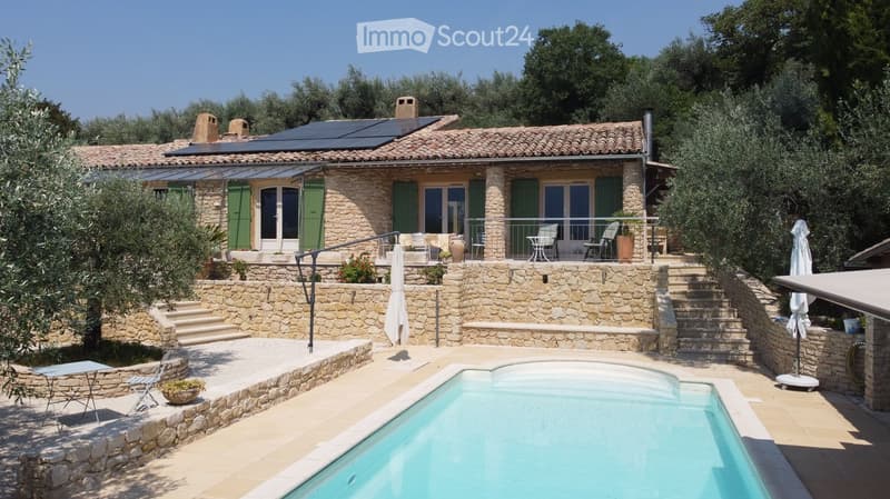 A stone house with a tiled roof, surrounded by greenery, features a swimming pool, outdoor seating, and steps leading up to the house.