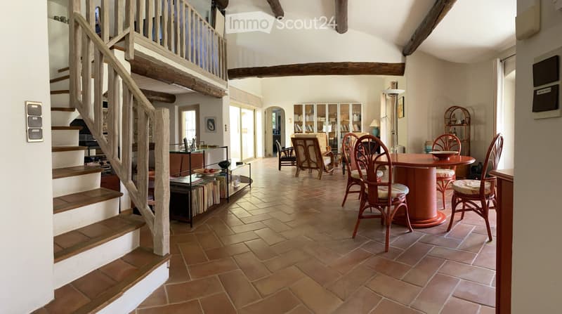 Spacious living area with terracotta tiled floor, wooden staircase leading to an upper level, round wooden table with chairs, bookshelf, and glass display shelf.