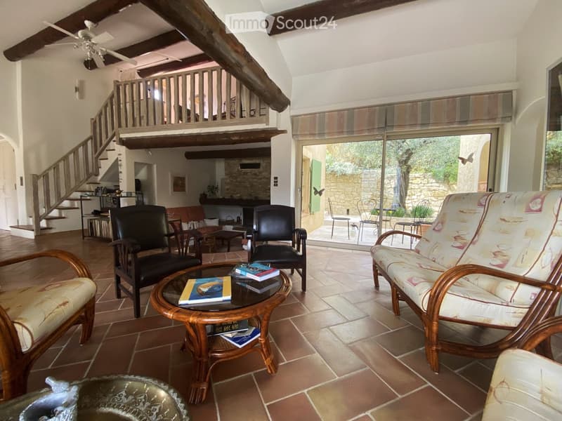 Living room with exposed wooden beams, tiled floor, and large glass doors leading to an outdoor area. Features a sofa, chairs, a coffee table with books, and a ceiling fan.