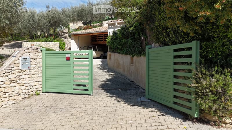 A gated entrance leads to a driveway with a garage and parking area, surrounded by greenery and stone walls.