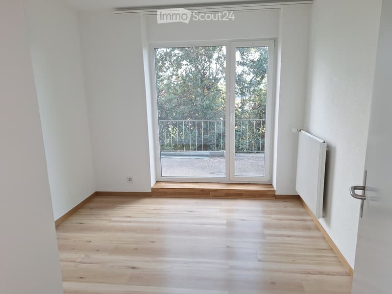 Empty room with light wooden flooring, white walls, and large windows leading to a balcony. A radiator is visible on one wall.