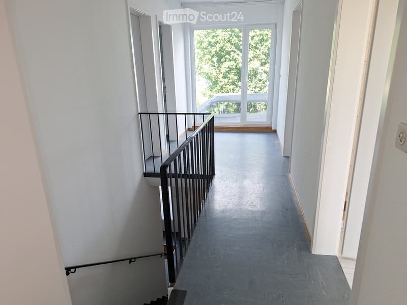 Empty hallway with grey tiled floor, white walls, and a staircase with a black metal railing leading to an upper level. Large windows at the end allow natural light.