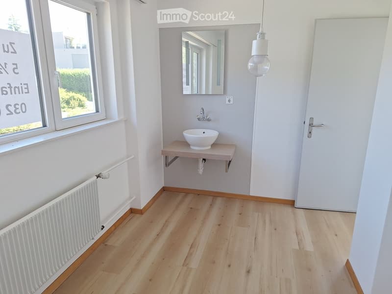 Modern bathroom with light wooden flooring, white walls, a sink with a mirror above, and a radiator near the window.