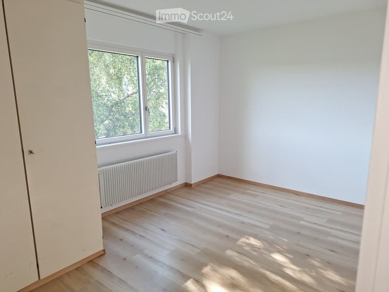 Empty room with light wooden flooring, white walls, and a window with a radiator below it.