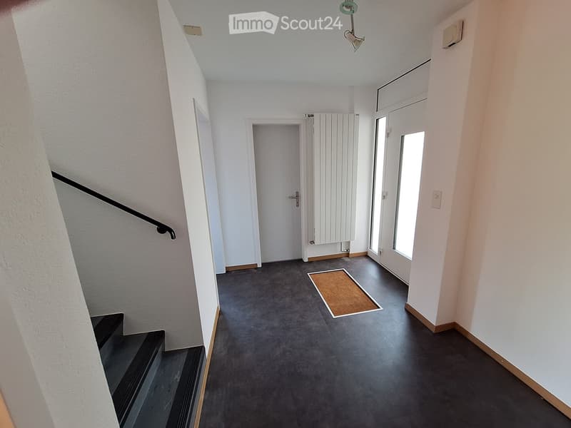 Empty hallway with grey carpeted floor, white walls, a staircase on the left, a door at the end, and a small doormat in the center.