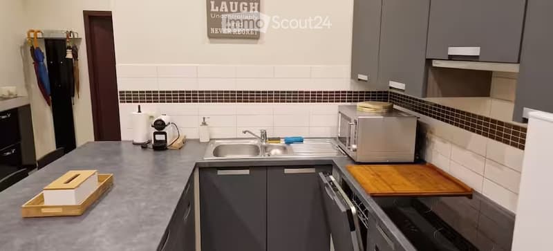 Modern kitchen with grey cabinets, stainless steel appliances, and a grey countertop. Features a sink, microwave, oven, dishwasher, coffee maker, and a wooden cutting board.