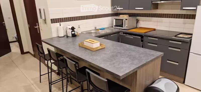 Modern kitchen with grey cabinets, a central island with a granite countertop, four stools, microwave, dishwasher, and a coffee maker on the counter.