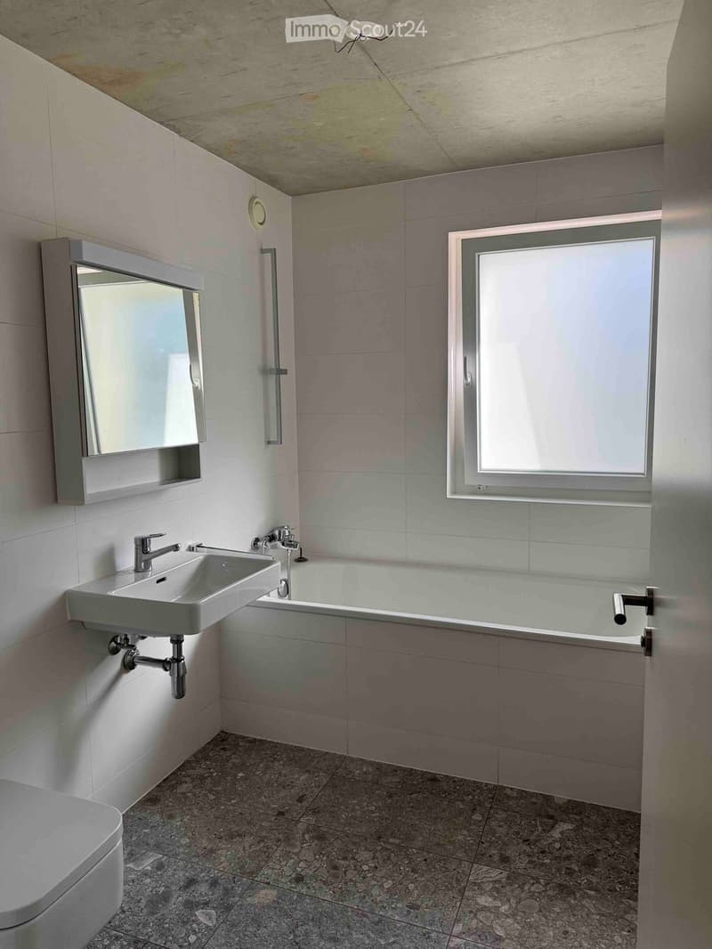 Modern bathroom with a white sink, bathtub, mirror, and toilet. Light-colored tiles and a window provide natural light.