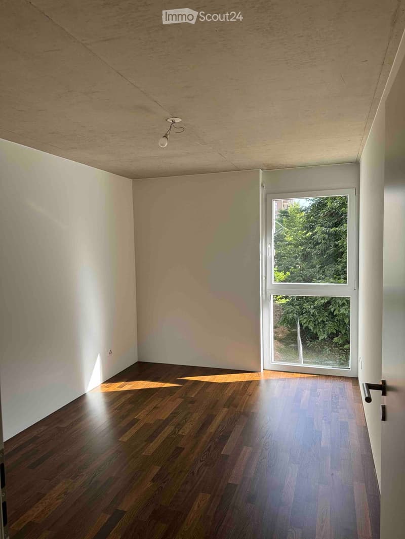 Empty room with wooden flooring, white walls, and a large window showing greenery outside. No furniture or equipment present.