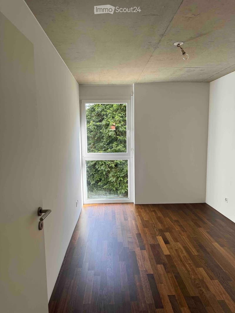 Empty hallway with wooden flooring, white walls, and a large window showing greenery outside. The ceiling has a single light fixture.
