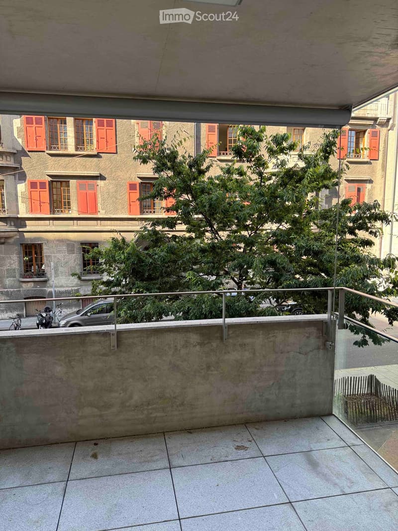 Balcony with tiled floor, metal railing, overlooking a tree-lined street and historic buildings with red shutters.