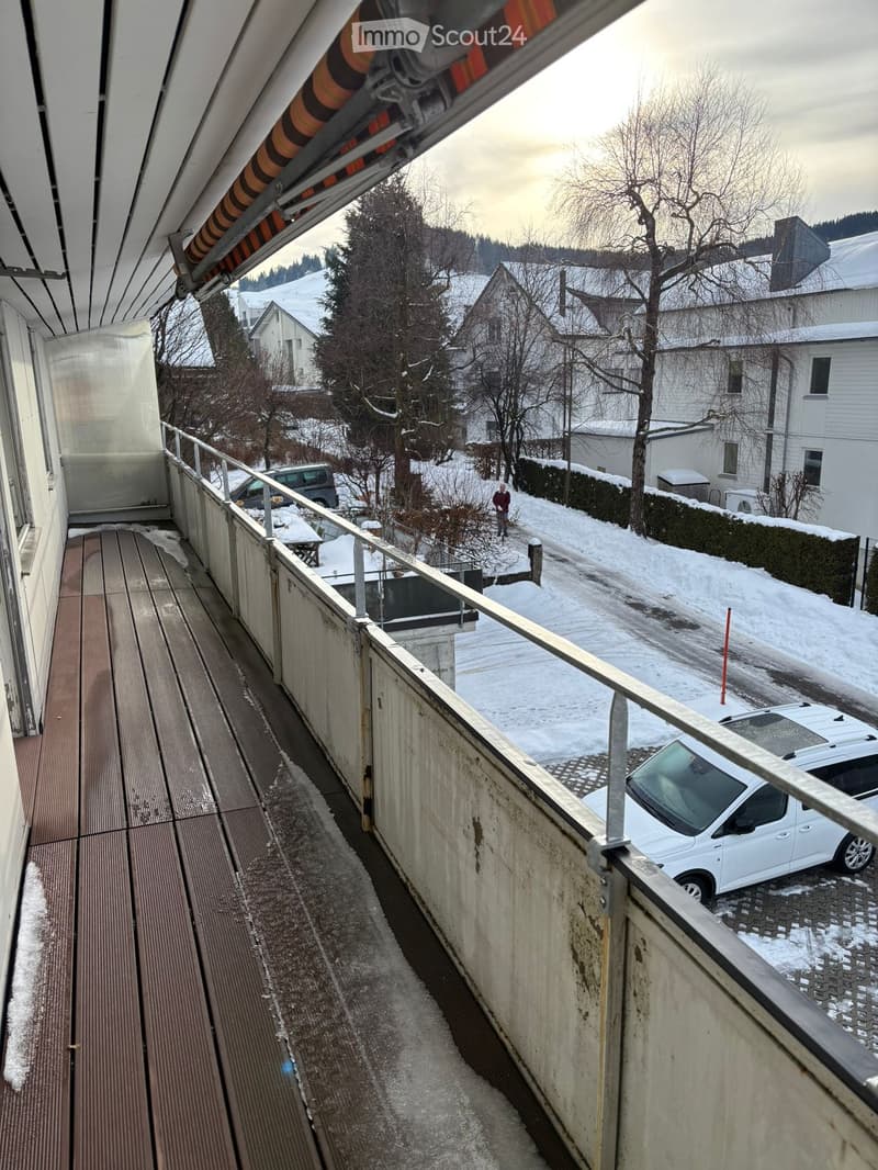 A balcony with wooden floor, metal railings, and retractable sun shade. View from balcony of snowy road, parked cars, and a person walking.