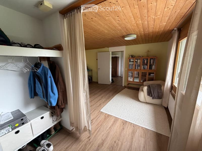 A cozy, well-lit room with wooden flooring, a small sofa with a throw, a bookcase, a curtained doorway, and a wardrobe with hanging clothes and shoes.