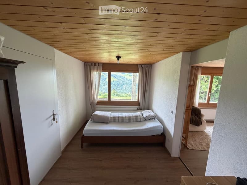 Cozy bedroom with wooden ceiling, large window with curtains, single bed, and a small chair.