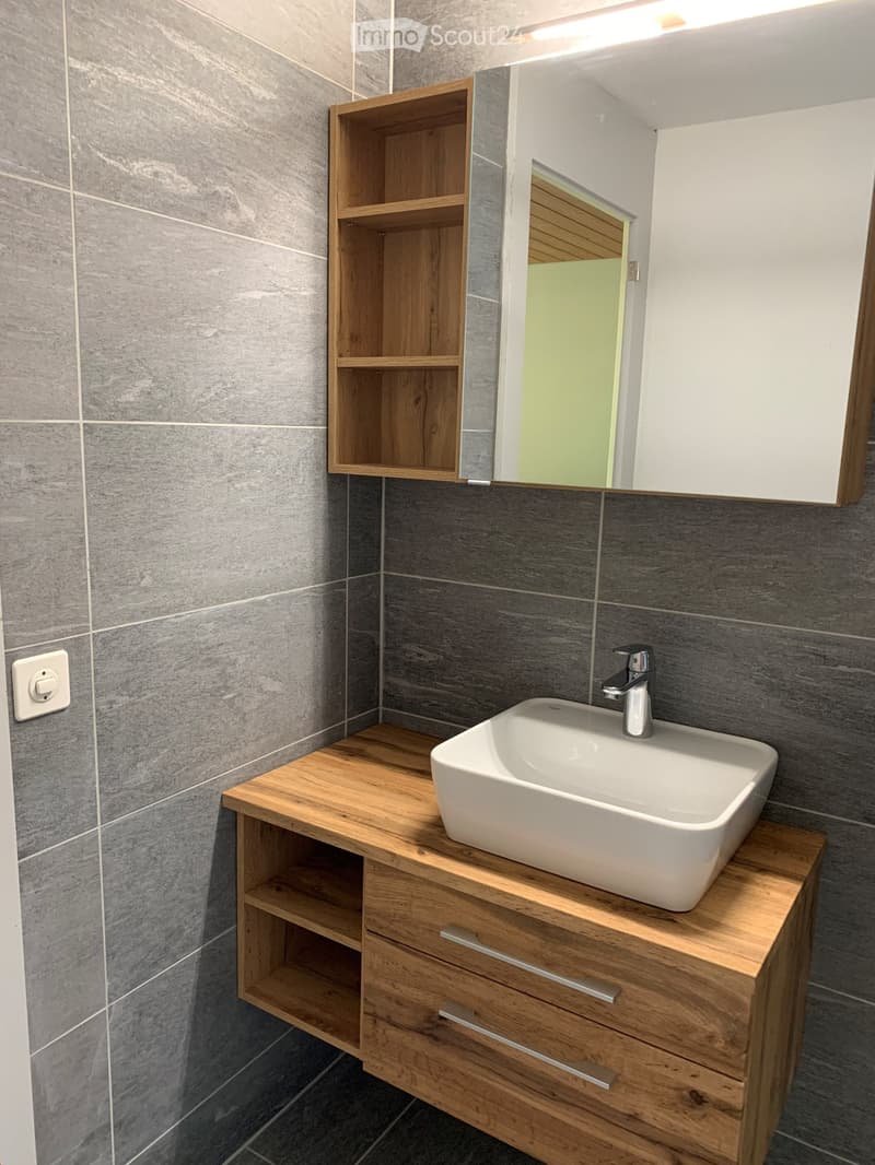 Modern bathroom with a white sink, wooden cabinet, and built-in wooden shelf. Grey tiled walls and a mirrored cabinet above the sink.