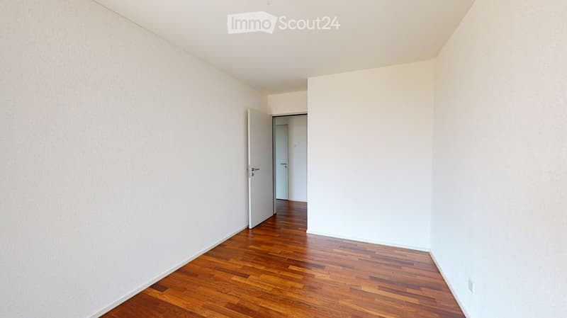 Empty room with white walls and wooden flooring, featuring an open doorway leading to another room.