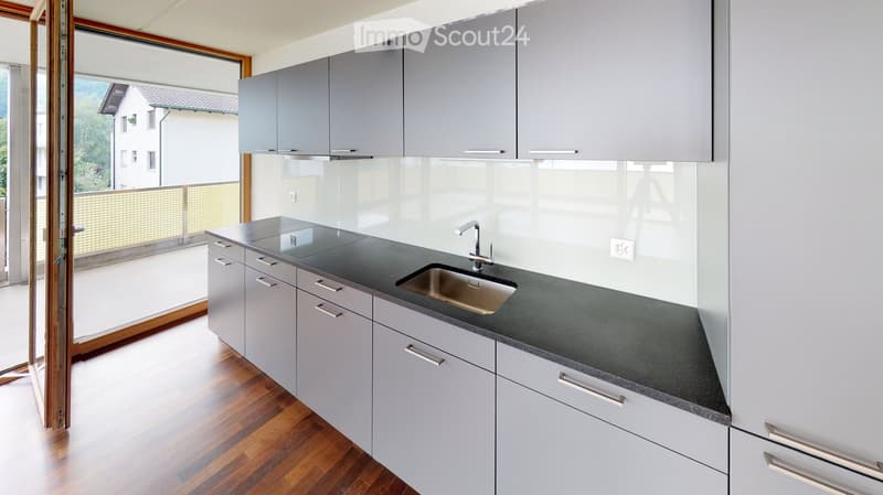 Modern kitchen with grey cabinets, black countertop, stainless steel sink, and wooden flooring.