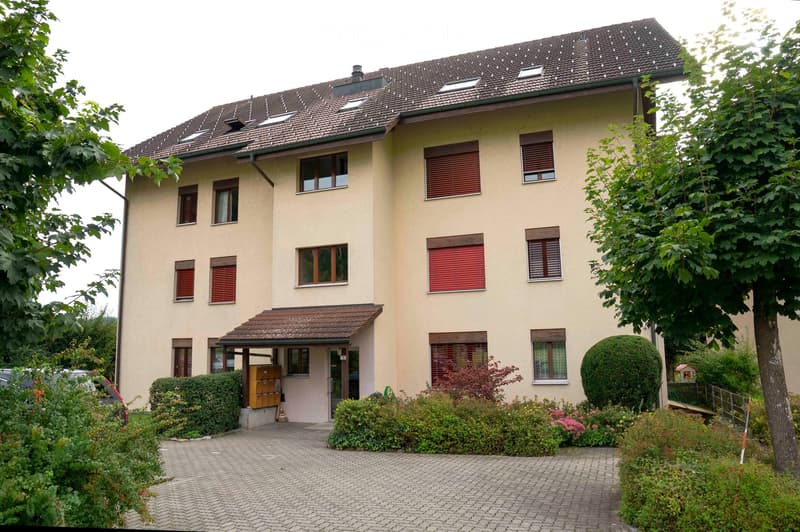 A three-story residential building with several windows, some of which have red shutters. The building has a brown roof, a small awning over the entrance, and a paved driveway. There are trees and bushes in front of the building, and a small garden area w