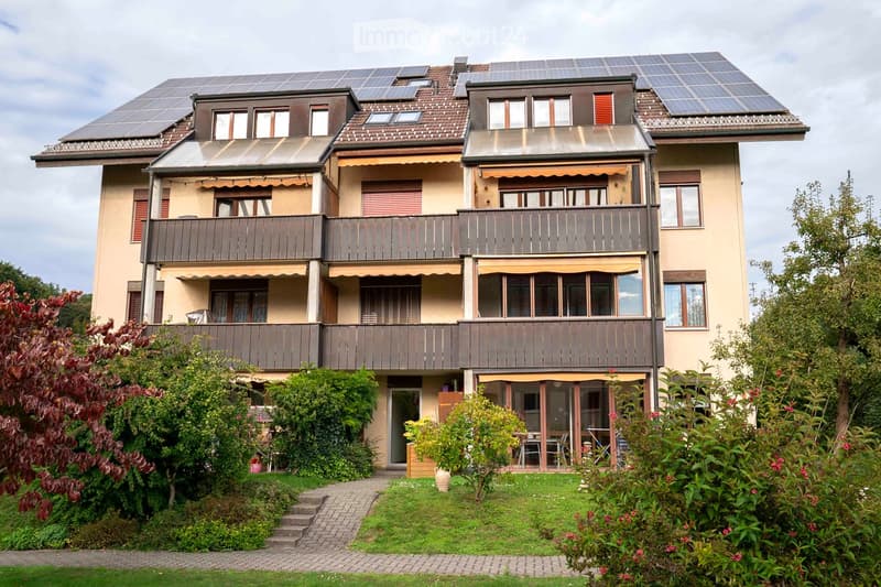 Three story apartment building with solar panels on the roof, multiple balconies, pathway, and garden