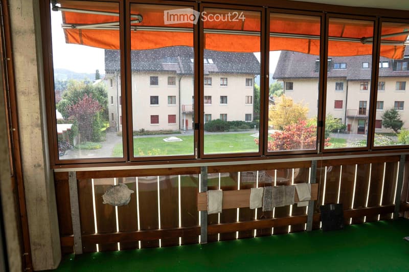 Carpeted floor, wooden slats with drying clothes, sliding glass doors, and balcony railings, outside view of a garden and a building
