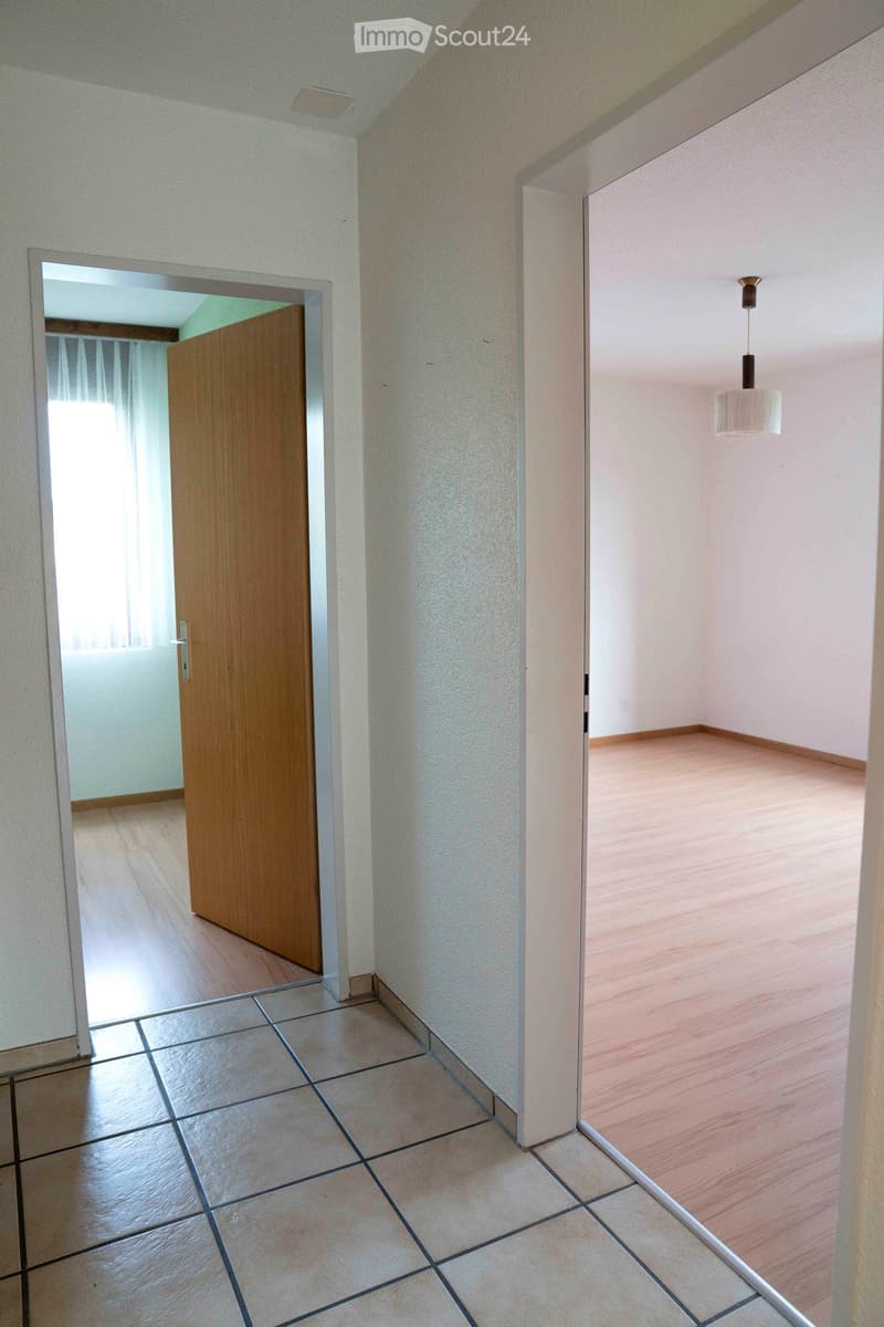 tiled floor, wooden door, white walls, open door leading to another room, natural light from window, wooden flooring in another room, ceiling light fixture