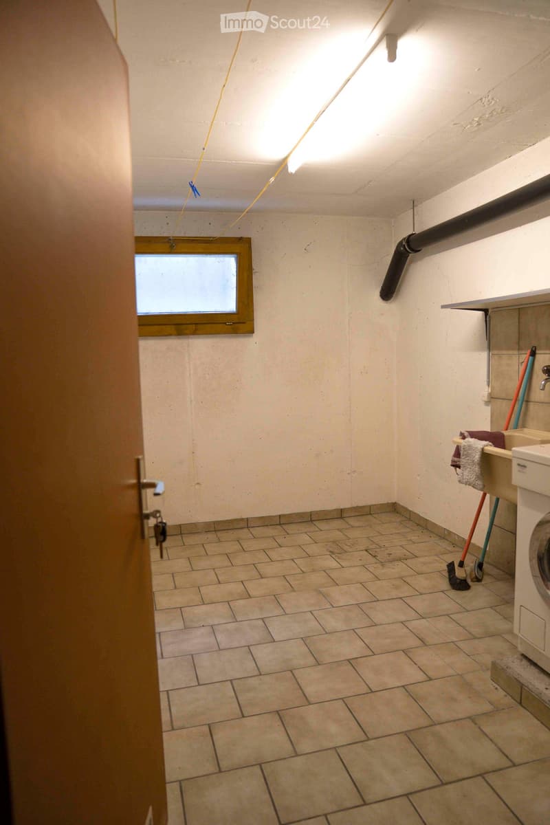 Small room with a small window, tiled floor, laundry washing machine, sink, mop and brush.