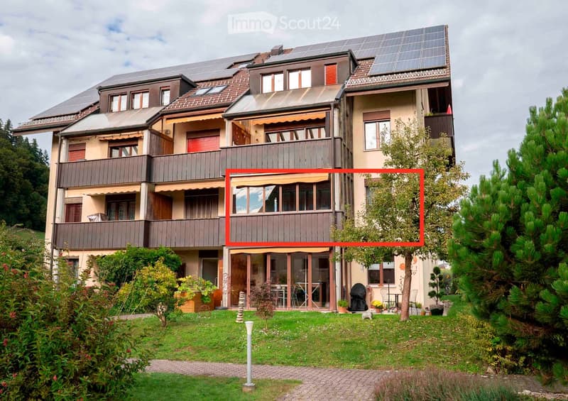 Multi-story apartment building with solar panels on the roof, balconies on each floor, and a ground floor entrance highlighted by a red box.