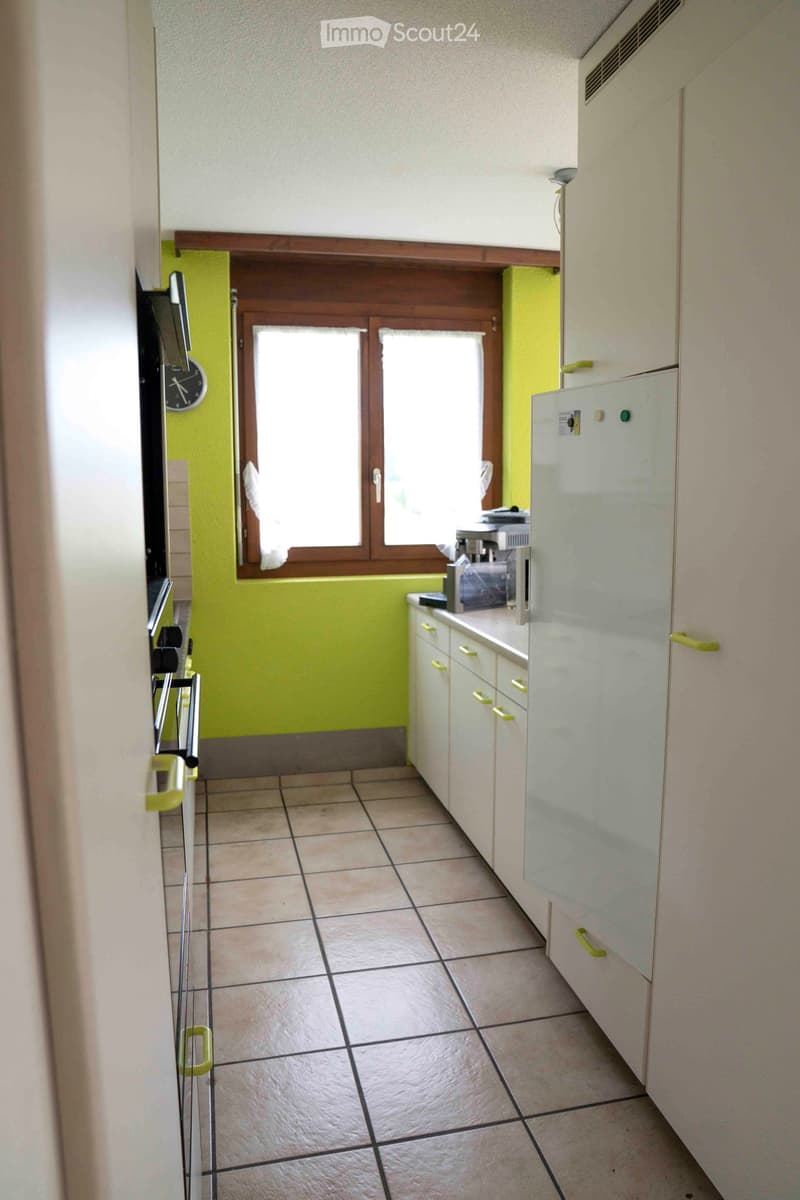 Kitchen: green wall, white tiled floor, white kitchen cabinets, white refrigerator, window with white curtains, oven, countertop with various appliances