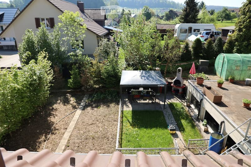 Garden with a small lawn, patio area with a table and chairs, barbecue, potted plants, and a greenhouse. The area is surrounded by trees and includes a house in the background.