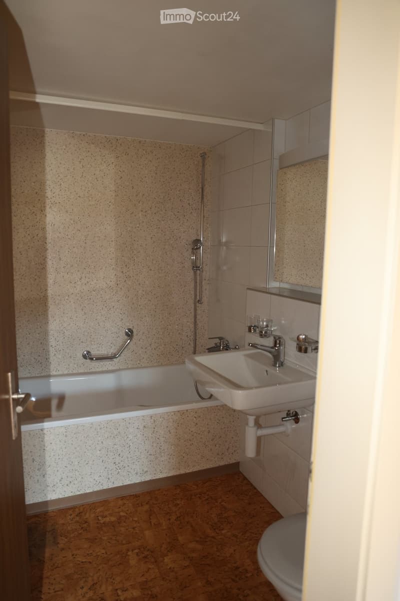 Modern bathroom with a bathtub, sink, toilet, and a handrail for accessibility. The walls are tiled with a speckled pattern, and the floor is made of brown tiles.