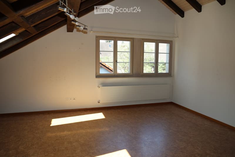 Empty attic room with wooden slanted ceiling, exposed beams, large window, radiator, and wooden floor.