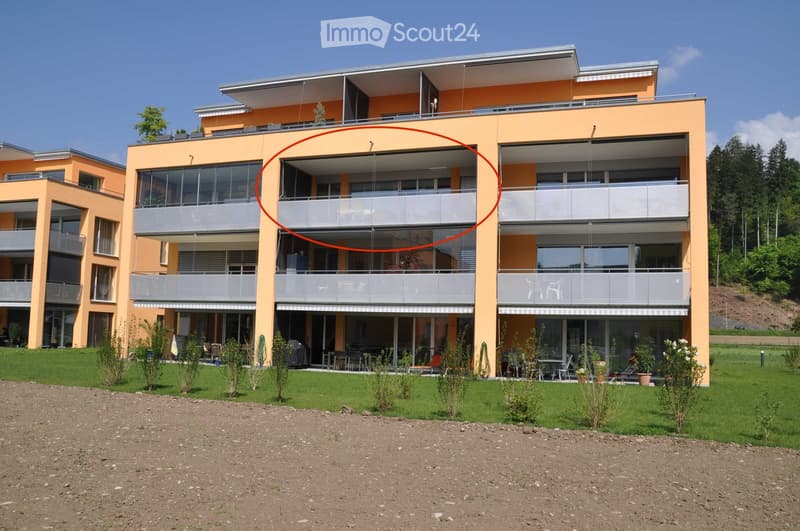 Modern two-story apartment building with orange facade, large windows, and multiple balconies. The ground floor features a well-maintained lawn, outdoor seating, and small plants.