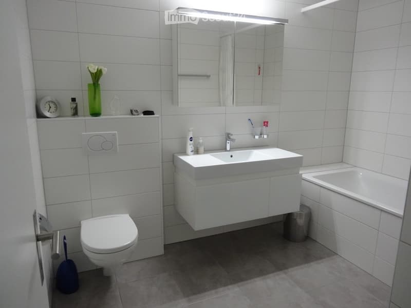 Modern bathroom with white tiles, a toilet, a sink with a mirror above it, a bathtub, and a small shelf with a vase of flowers and a clock.