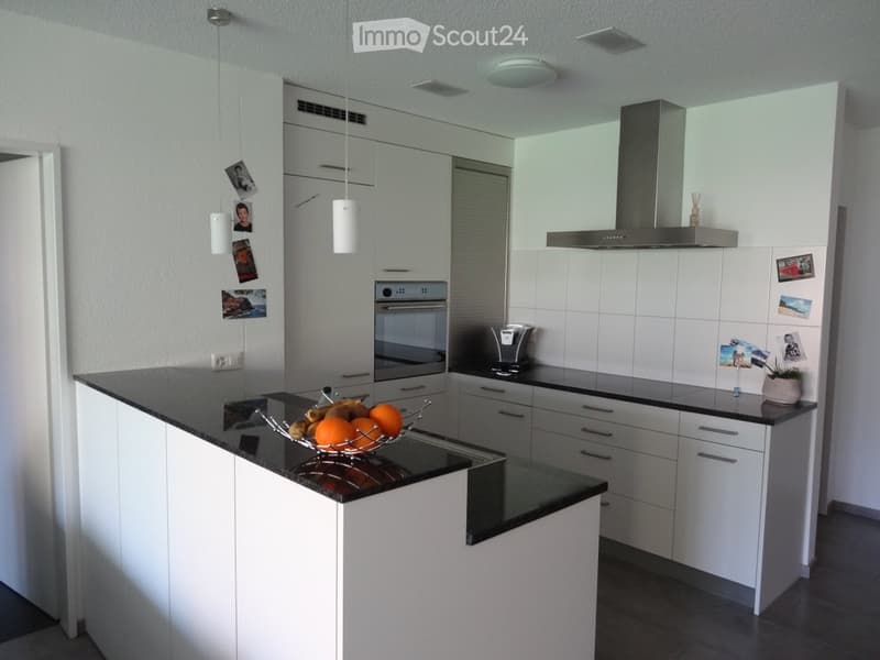Modern kitchen with white cabinets, black countertops, and stainless steel appliances, including an oven and a coffee maker. A fruit bowl with oranges and bananas is placed on the counter.