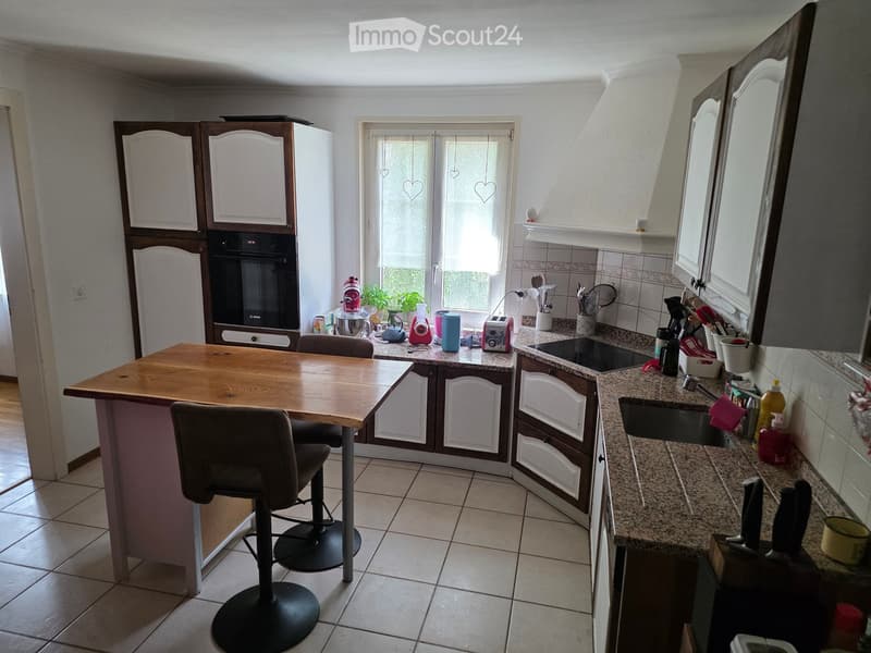 A modern kitchen with a central island, granite countertops, and various appliances including a refrigerator, oven, and sink. The island features a wooden table with two chairs, and the countertops are cluttered with kitchen items such as a toaster, cups,