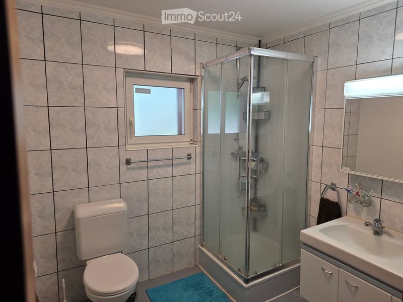 Modern bathroom with tiled walls, a toilet, a sink with a mirror, a glass-enclosed shower, and a towel on the floor.