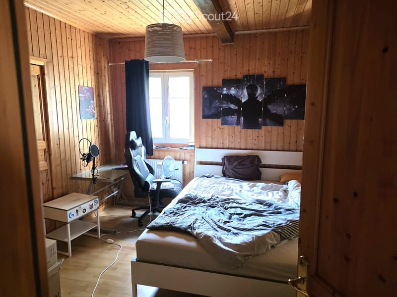 Cozy wooden bedroom with a bed, desk, chair, fan, and artwork on the walls.