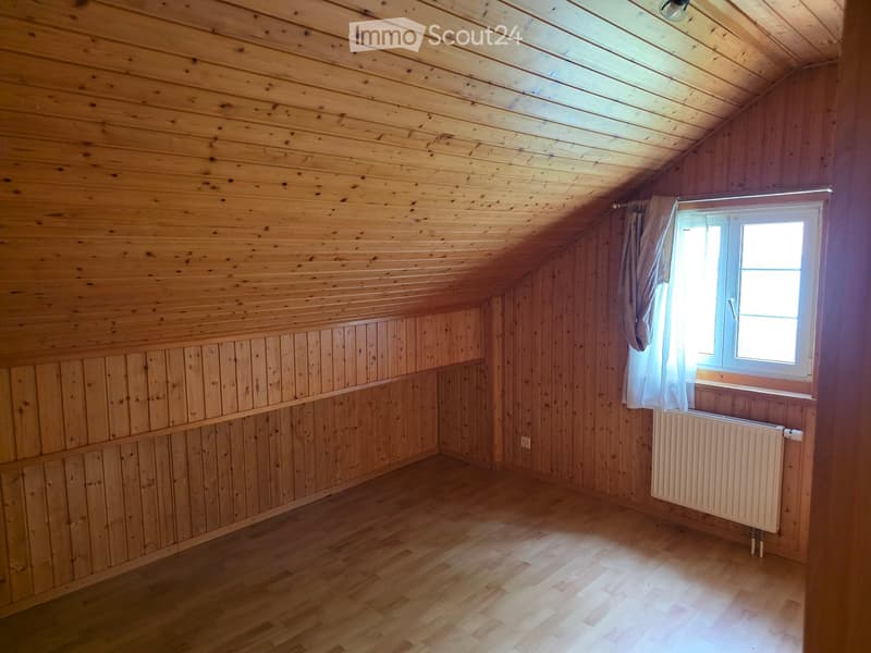 Empty attic room with wooden walls, ceiling, and floor, featuring a window with a curtain and a radiator below it.
