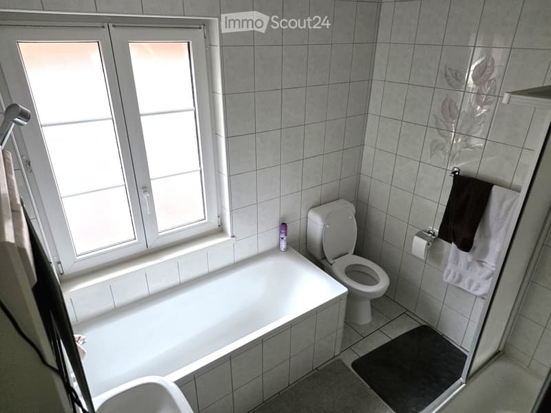 Modern bathroom with a bathtub, toilet, and a glass shower enclosure. The room features tiled walls and a window with frosted glass panes.