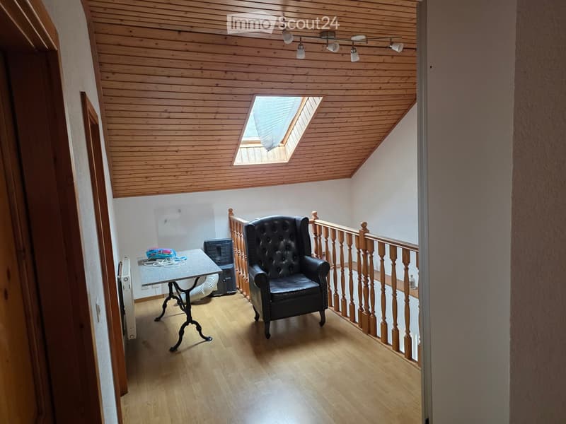 A well-lit room with wooden flooring, a black leather armchair, a small table, and a radiator. The room features a wooden staircase railing and a skylight in the ceiling.