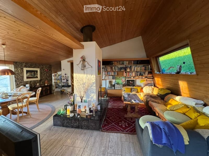 Open-concept living and dining area with wooden ceilings, a stone accent wall, a large sofa with colorful cushions, a coffee table, a dining table with chairs, and a bookshelf filled with books.