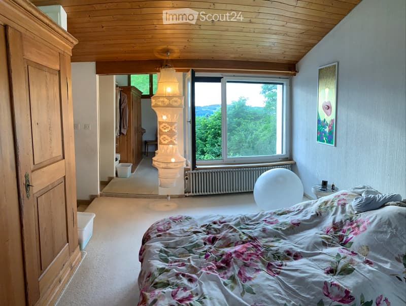 Bright bedroom with wooden ceiling, large window offering a scenic view, bed with floral bedding, wardrobe, small table, and a hanging lamp.