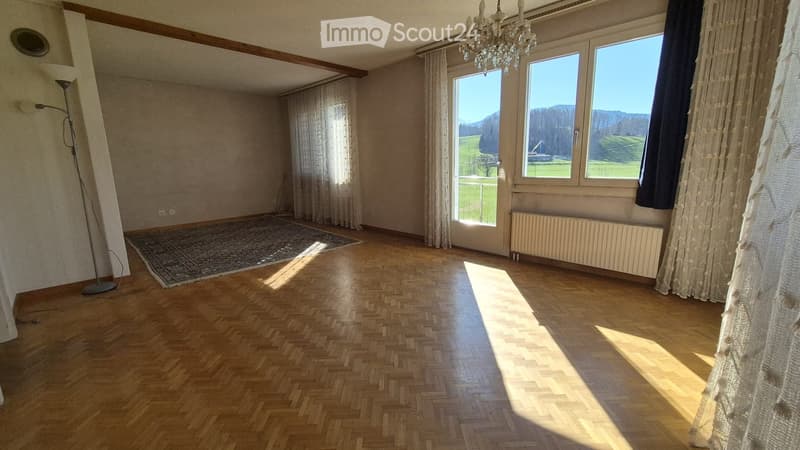 Empty room with wooden herringbone flooring, large windows with curtains, a chandelier, and a radiator. The room has a view of a green landscape outside.