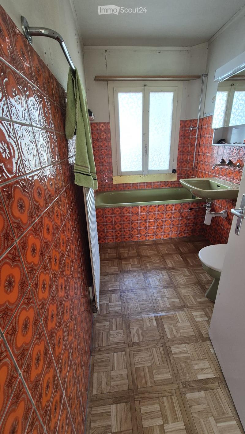 Bathroom with wooden flooring, orange and white tiled walls, green bathtub, sink, and a towel hanging on the wall.