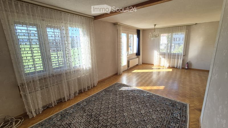 Empty room with wooden flooring, large windows with sheer curtains, and a patterned rug on the floor.